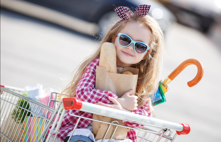 Little girl,with thick blonde long hair,dressed in a plaid shirt and denim shorts,on his head a black-and-red bow,wears sun glasses,sitting in the shopping cart on wheels near a large supermarket Stylishe Kinder